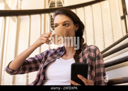 Kaffee trinken und Smartphone benutzen, Frau, die sich zu Hause auf der Treppe entspannt Stockfoto