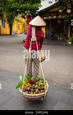 Vietnamesische Frau, die Körbe mit Früchten trägt. Die örtliche Verkäuferin verkauft Obst, das sie in Körben in der Altstadt von Hoi an, Vietnam, trägt. Vietnamesische Straße Stockfoto