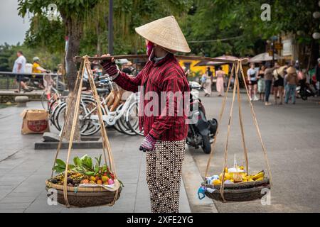 Vietnamesische Frau, die Körbe mit Früchten trägt. Die örtliche Verkäuferin verkauft Obst, das sie in Körben in der Altstadt von Hoi an, Vietnam, trägt. Vietnamesische Straße Stockfoto