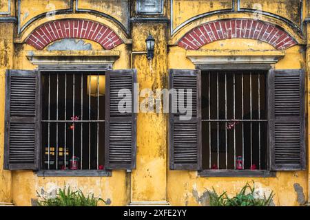 Zwei alte Fenster mit Holzrahmen und Rollläden, ein Fragment der Fassade eines alten Gebäudes in Hoi an Vietnam. Gelb lackierte Hauswand mit Rissen Stockfoto
