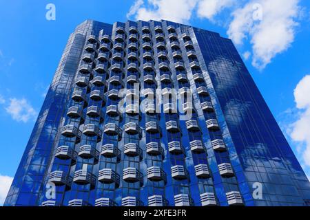Blick nach oben auf einen hohen Wolkenkratzer mit sich wiederholenden Balkonen und einer Glasfassade, die den blauen Himmel und die Wolken reflektiert. Stockfoto