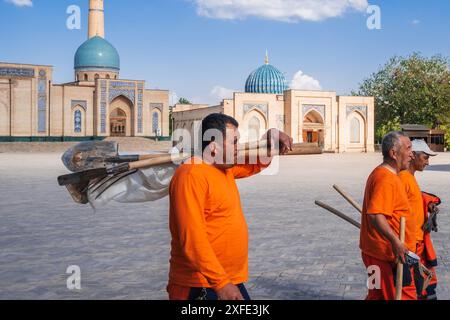 Usbekische männliche Hausmeister in orangefarbenen Uniformen mit Schaufeln auf der Straße im Frühling auf dem Platz des Hazrati Imam Architekturkomplexes. Taschkent, Usbekistan - 17. April 2024 Stockfoto