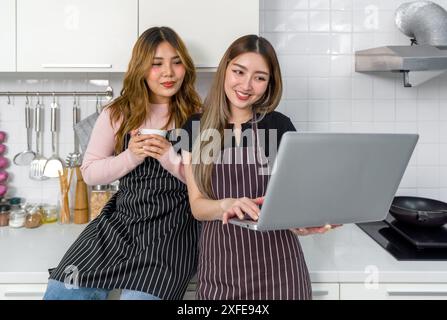 Zwei Frauen mit gestreifter Schürze lesen oder mit Laptop-Bildschirm in einer modernen Küche interagieren. Kochutensilien stehen auf der Arbeitsplatte. Stockfoto