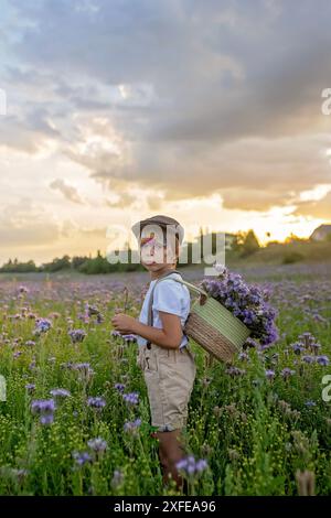 Wunderschönes Schulkind, Junge mit bemaltem Gesicht in einem Blumenfeld bei Sonnenuntergang, Korb mit Blumen und alten Vintage Koffer, Sommer Stockfoto