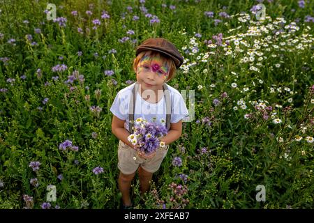 Wunderschönes Schulkind, Junge mit bemaltem Gesicht in einem Blumenfeld bei Sonnenuntergang, Korb mit Blumen und alten Vintage Koffer, Sommer Stockfoto