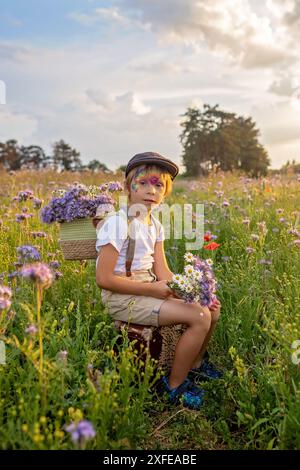 Wunderschönes Schulkind, Junge mit bemaltem Gesicht in einem Blumenfeld bei Sonnenuntergang, Korb mit Blumen und alten Vintage Koffer, Sommer Stockfoto