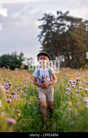 Wunderschönes Schulkind, Junge mit bemaltem Gesicht in einem Blumenfeld bei Sonnenuntergang, Korb mit Blumen und alten Vintage Koffer, Sommer Stockfoto