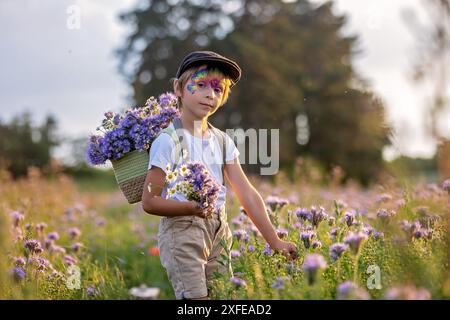 Wunderschönes Schulkind, Junge mit bemaltem Gesicht in einem Blumenfeld bei Sonnenuntergang, Korb mit Blumen und alten Vintage Koffer, Sommer Stockfoto