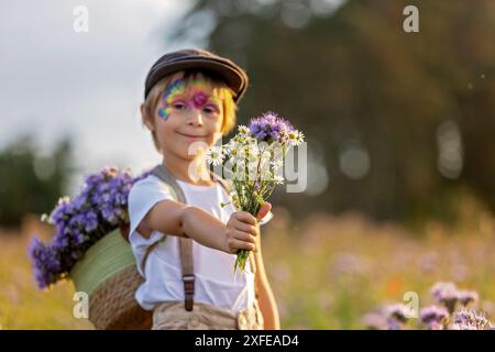 Wunderschönes Schulkind, Junge mit bemaltem Gesicht in einem Blumenfeld bei Sonnenuntergang, Korb mit Blumen und alten Vintage Koffer, Sommer Stockfoto