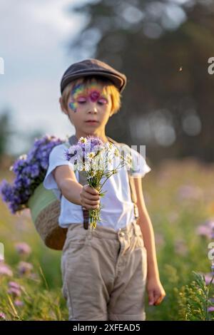 Wunderschönes Schulkind, Junge mit bemaltem Gesicht in einem Blumenfeld bei Sonnenuntergang, Korb mit Blumen und alten Vintage Koffer, Sommer Stockfoto