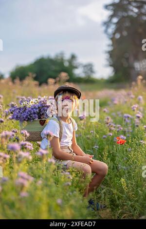 Wunderschönes Schulkind, Junge mit bemaltem Gesicht in einem Blumenfeld bei Sonnenuntergang, Korb mit Blumen und alten Vintage Koffer, Sommer Stockfoto