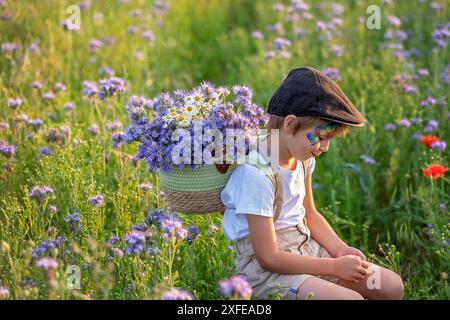 Wunderschönes Schulkind, Junge mit bemaltem Gesicht in einem Blumenfeld bei Sonnenuntergang, Korb mit Blumen und alten Vintage Koffer, Sommer Stockfoto