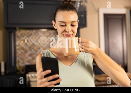 Smartphone halten und Kaffee trinken, Frau genießt den Morgen in der Küche Stockfoto