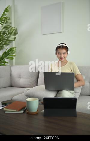 Lächelnde junge asiatische Frau trägt kabellose Kopfhörer, die auf dem Sofa liegen und einen Laptop benutzen Stockfoto