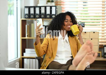 Sorglose afroamerikanische Geschäftsfrau mit überkreuzten Beinen auf dem Tisch, genießen Sie morgens Kaffee und telefonieren Stockfoto