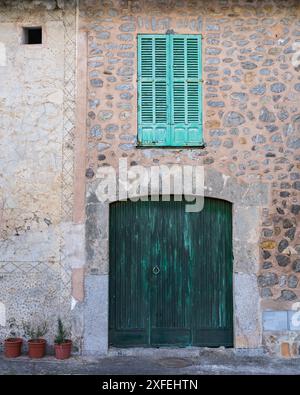 Eine alte grüne Holztür mit einem eisernen Griff in einer Steinmauer, oben ein Fenster mit Holzläden. Stockfoto