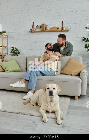 Ein bärtiges schwules Paar genießt eine schöne Zeit mit ihrem Labrador auf einer bequemen Couch in ihrem Wohnzimmer. Stockfoto