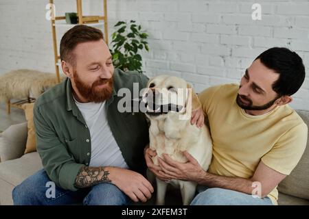 Bärtiges Schwulenpaar, das auf einer Couch sitzt und freudig ihren Labrador in einer gemütlichen Wohnzimmerumgebung hält und streichelt. Stockfoto