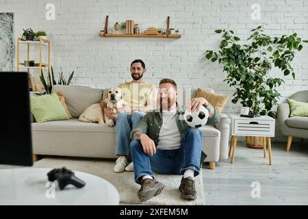 Ein bärtiger Mann sitzt auf einer Couch, hält einen Fußball, tief in Gedanken, und verbringt schöne Zeit im Wohnzimmer. Stockfoto