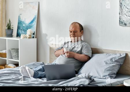 Ein Mann mit Inklusivität sitzt im Bett, hält eine Tasse Kaffee und schaut auf seinen Laptop. Stockfoto