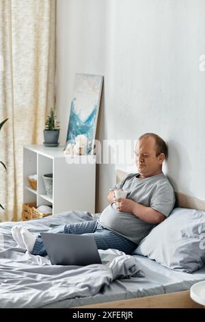 Ein Mann mit Inklusivität sitzt im Bett, hält eine Tasse Kaffee und benutzt einen Laptop. Stockfoto
