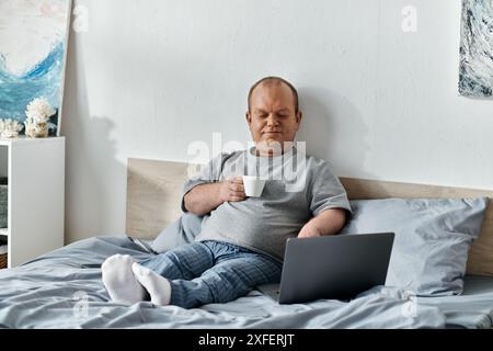 Ein Mann mit Inklusivität sitzt im Bett und genießt eine Tasse Kaffee, während er seinen Laptop benutzt. Stockfoto