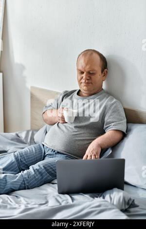 Ein Mann mit Inklusivität sitzt im Bett und genießt eine Tasse Kaffee, während er an seinem Laptop arbeitet. Stockfoto
