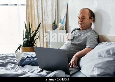 Ein Mann mit Inklusivität im Schlafanzug sitzt im Bett mit einem Laptop und einer Tasse Kaffee. Stockfoto