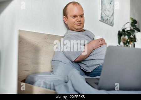 Ein Mann mit Inklusivität sitzt im Bett, genießt eine Tasse Kaffee und arbeitet an seinem Laptop. Stockfoto
