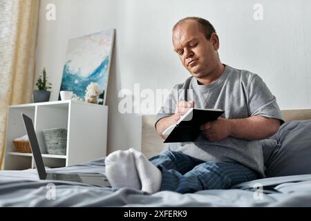 Ein Mann mit Inklusivität sitzt auf seinem Bett, schreibt in ein Notizbuch, mit einem Laptop neben ihm. Stockfoto