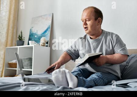 Ein Mann mit Inklusivität sitzt im Bett, arbeitet an einem Laptop und hält ein Buch. Stockfoto