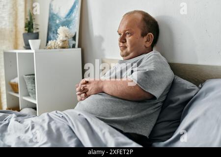 Ein Mann mit Inklusivität sitzt im Bett, verloren in Gedanken, während das Morgenlicht durch das Fenster filtert. Stockfoto