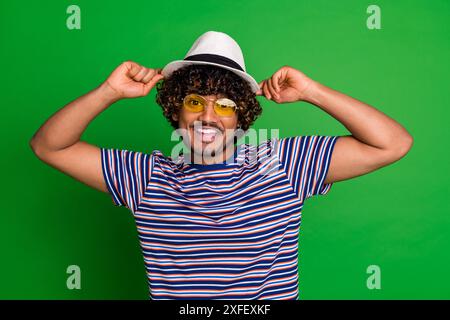 Porträt des jungen mexikanischen coolen lockigen lustigen Mannes, der Sonnenbrille und Sonnenhut-Schüler in gestreiftem T-Shirt auf grünem Farbhintergrund trägt Stockfoto