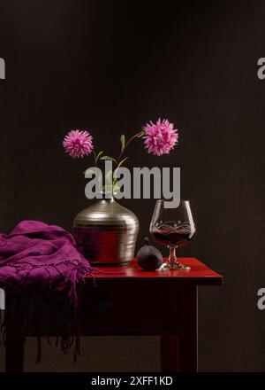 Zwei rosafarbene Blumen. Astern in einer grauen Vase auf einem roten Holztisch. In der Nähe liegt eine reife blaue Feige, ein dunkelrosa Schal. In der Nähe gibt es ein Glas Rotwein. Dunkles g Stockfoto