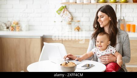 Frau, die am Laptop arbeitet, während sie ihr Baby in der Küche hält Stockfoto