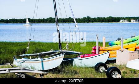 NORWALK, CT, USA - 2. JULI 2024: Zwei weiße und blaue Hobie Cat Segelboote sitzen am Ufer neben einem Stapel Kajaks Stockfoto