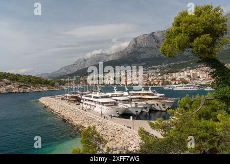 Touristenschiffe und Yachten befinden sich in der Bucht am Pier. Die Leute laufen am Damm entlang. Sonniger Sommertag. Hohe Klippen. Stadt Makarska Kroatien Stockfoto