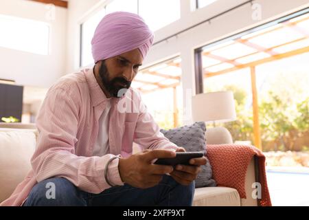 Mit Smartphone, Mann im Turban sitzt zu Hause auf der Couch und konzentriert sich Stockfoto