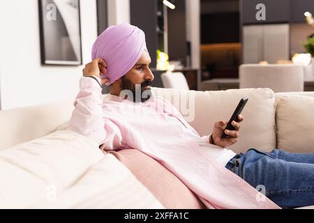 Entspannend auf der Couch, indischer Mann im Turban mit Smartphone zu Hause Stockfoto