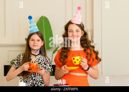 Zwei Mädchen, die Geburtstagshüte tragen und bunte Tassen halten, feiern drinnen. Stockfoto