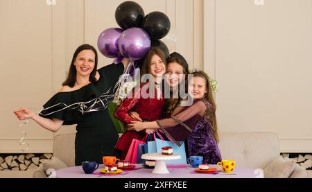 Teenager in rotem Spitzenkleid und zwei junge Mädchen, die sich umarmen, während eine Frau mit Ballons ansieht. Geburtstagsfeier Stockfoto