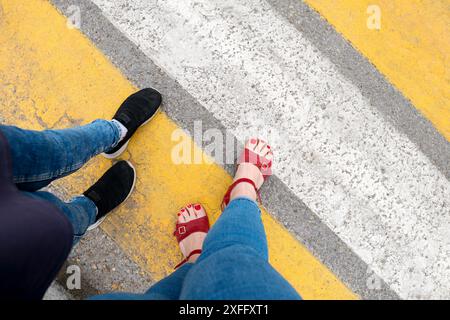 Füße in roten Sandalen mit roten Zehennägeln und schwarzen Schuhen auf einem gelb gestreiften Steg. Stockfoto