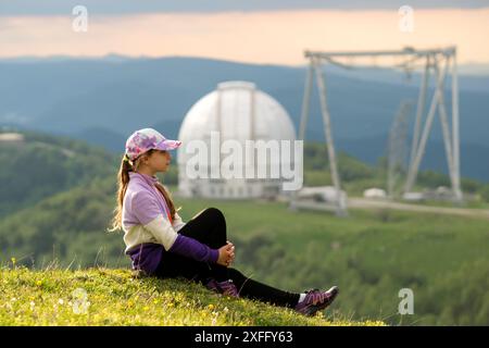 Mädchen, das auf einem grasbewachsenen Hügel sitzt, mit einem Observatorium im Hintergrund. Wanderer, die sich vor dem Hintergrund von BTA-6, grosser Altazimuth T ausruhen und posieren Stockfoto