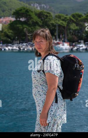 Eine Frau mittleren Alters mit einem Rucksack auf dem Rücken bereist die Welt und genießt neue Orte. Vor dem Hintergrund des blauen Meeres und einer touristischen Stadt in CR Stockfoto