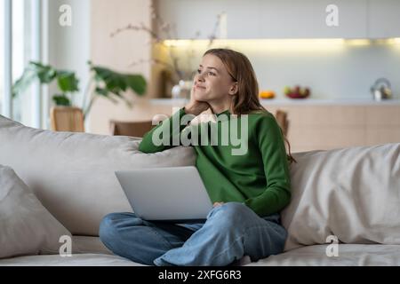 Nachdenkliche weibliche Fernarbeiterin, die sich auf dem Sofa mit Computer entspannt, vom Geschäft abgelenkt und nachgedacht hat. Stockfoto