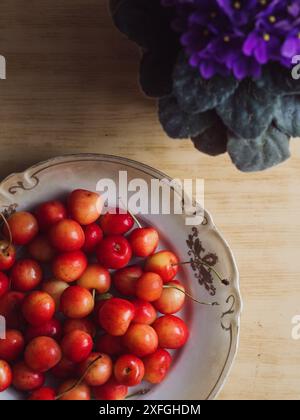 Süße Kirschen in Einem Beige Vintage Teller und ein violettes Violett in Bloom auf einem Holztisch. Vertikale Flache Lage Stockfoto