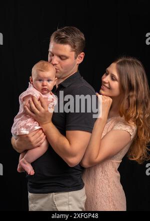 Eine junge Familie mit einem Baby in den Armen. Dad küsst das Neugeborene, Mom sieht zärtlich aus. Das Mädchen trägt leichte Kleidung, der Kerl trägt einen schwarzen T-sh Stockfoto