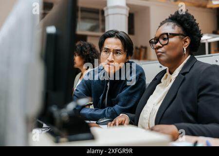 Ein konzentrierter Geschäftsmann sitzt mit einer Kollegin, während er im Büro den Computerbildschirm teilt Stockfoto