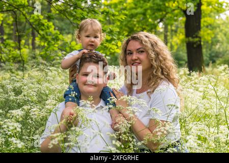 Mom mit zwei Söhnen im dicken Gras. Der ältere Bruder hält ein kleines Baby, ein Jahr alt, auf seinen Schultern. Glückliche Familie Stockfoto