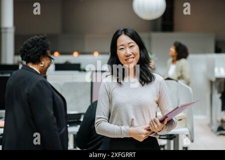 Porträt einer lächelnden Geschäftsfrau, die Dokumente hält, während sie am Arbeitsplatz steht Stockfoto